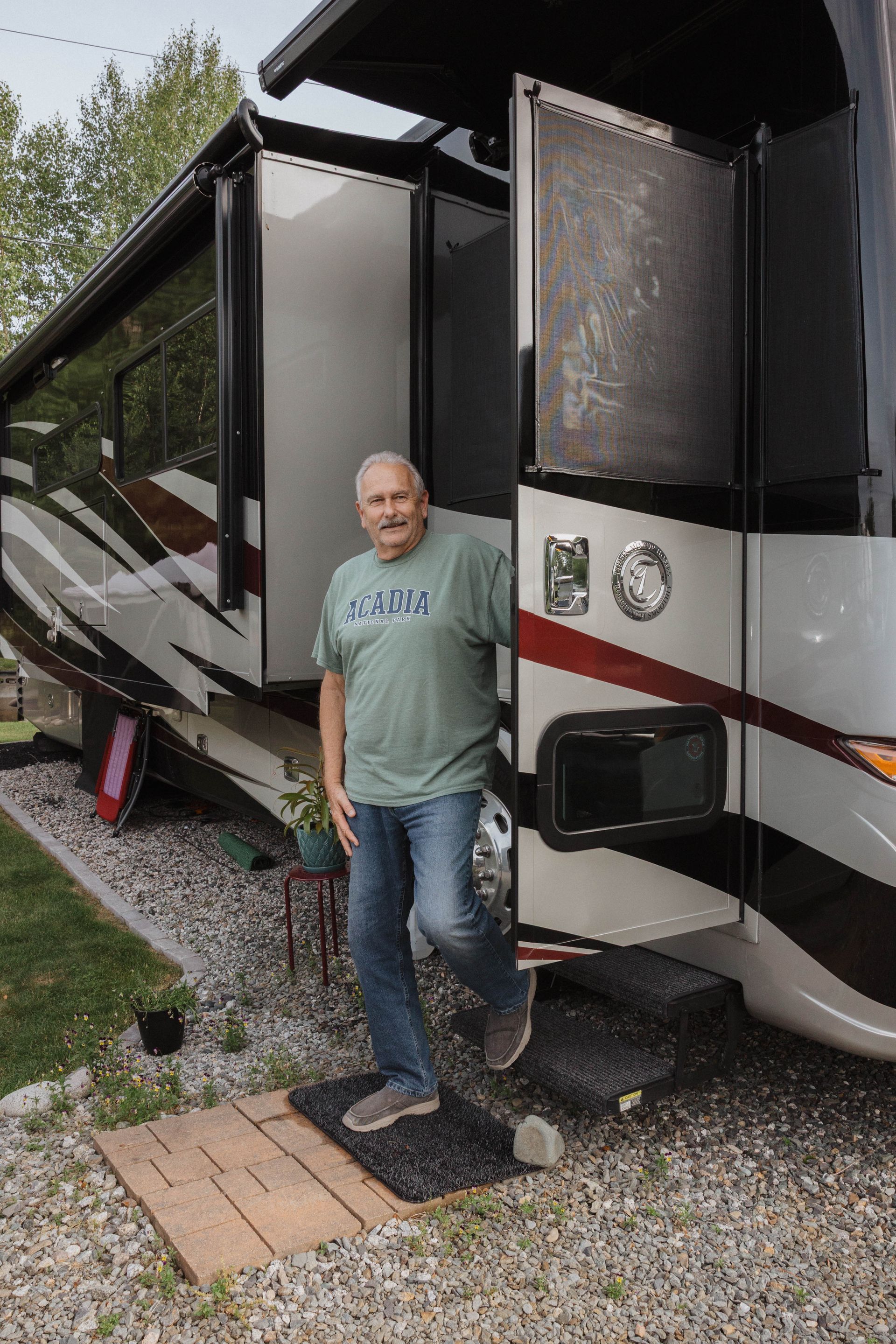 Man standing beside a large black and white RV with the door open on a gravel campsite.