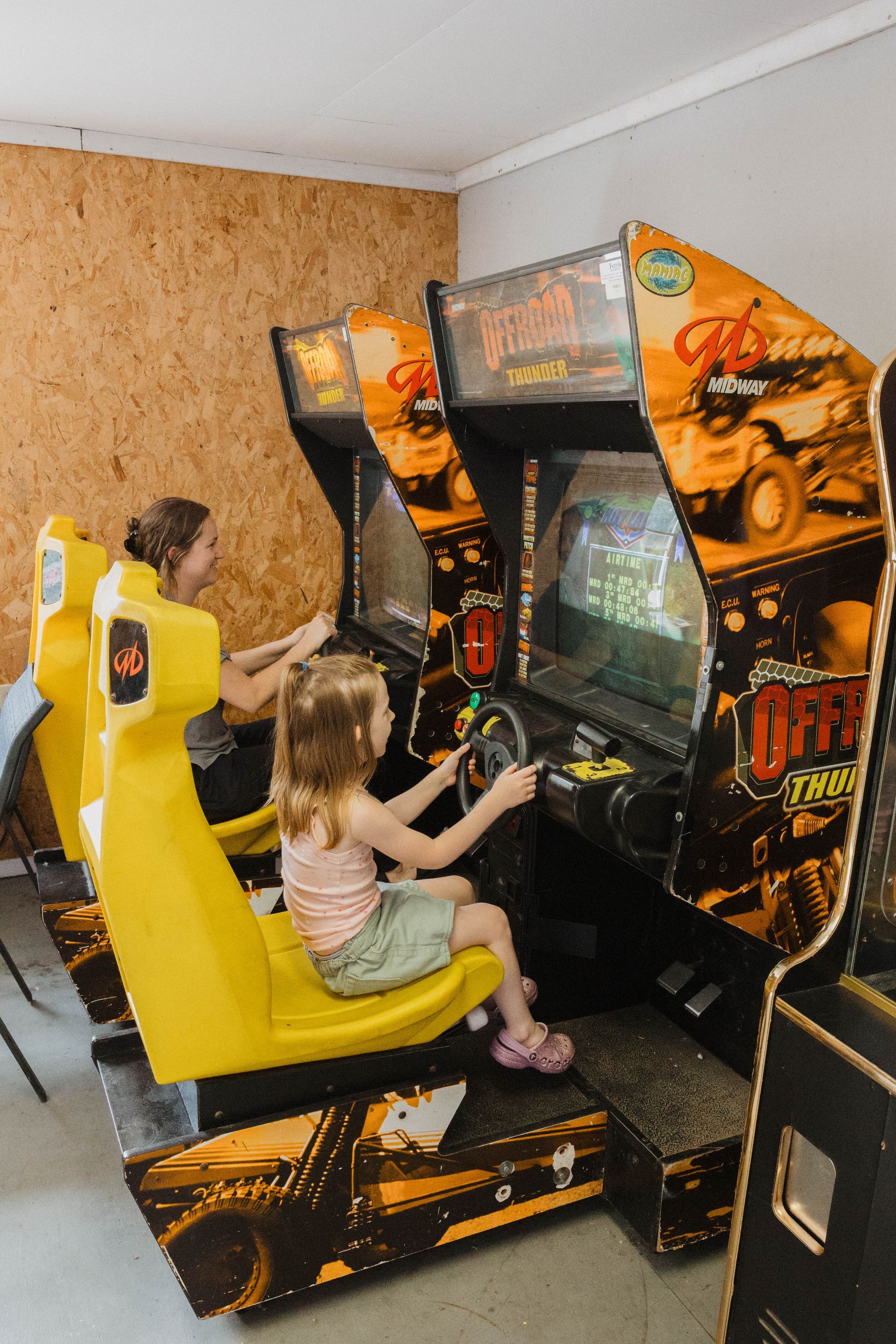 Child playing on a yellow arcade racing game in a room with wood-paneled wall and stacked machines