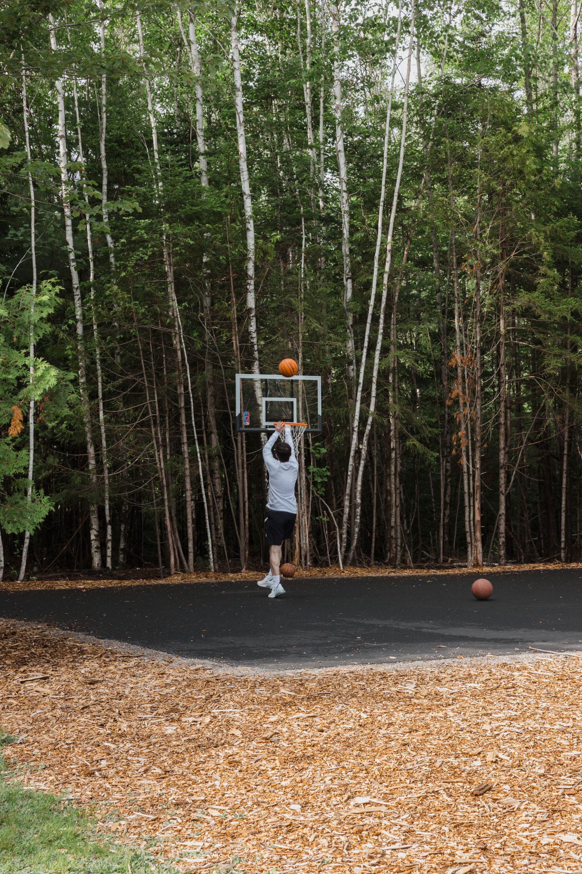 Person shooting a basketball outdoors near a birch forest, with an orange ball on the hoop