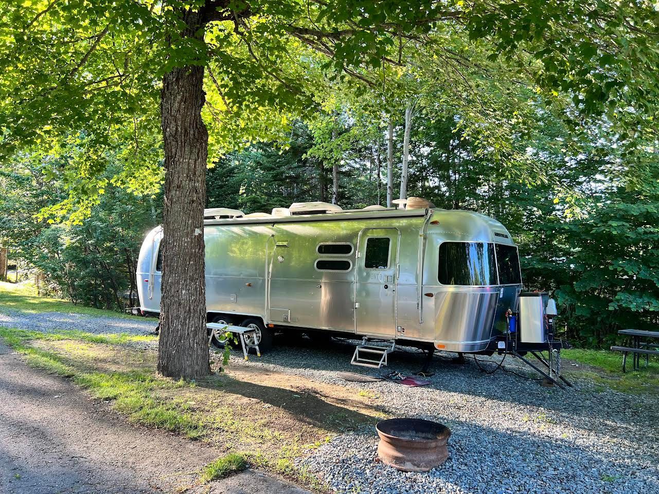Silver travel trailer parked under leafy trees at a campsite with a campfire ring in front