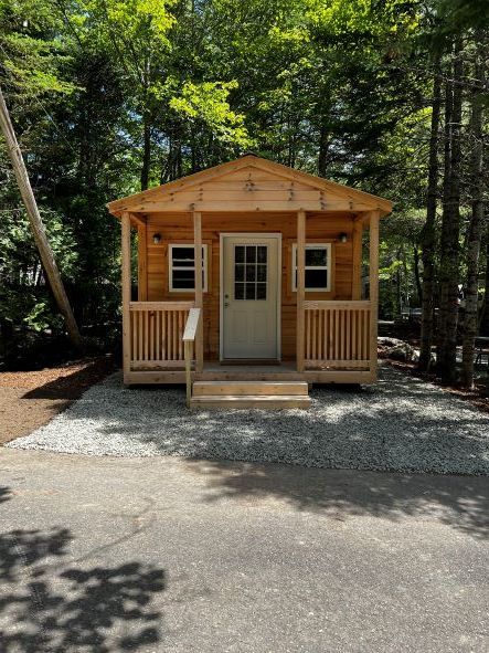 Small wooden cabin with a front porch and steps, surrounded by trees on a gravel lot.