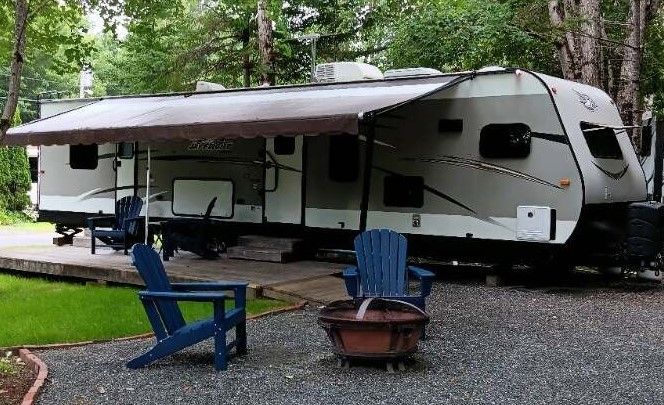 White travel trailer in a wooded campsite with blue chairs and a fire pit on gravel.