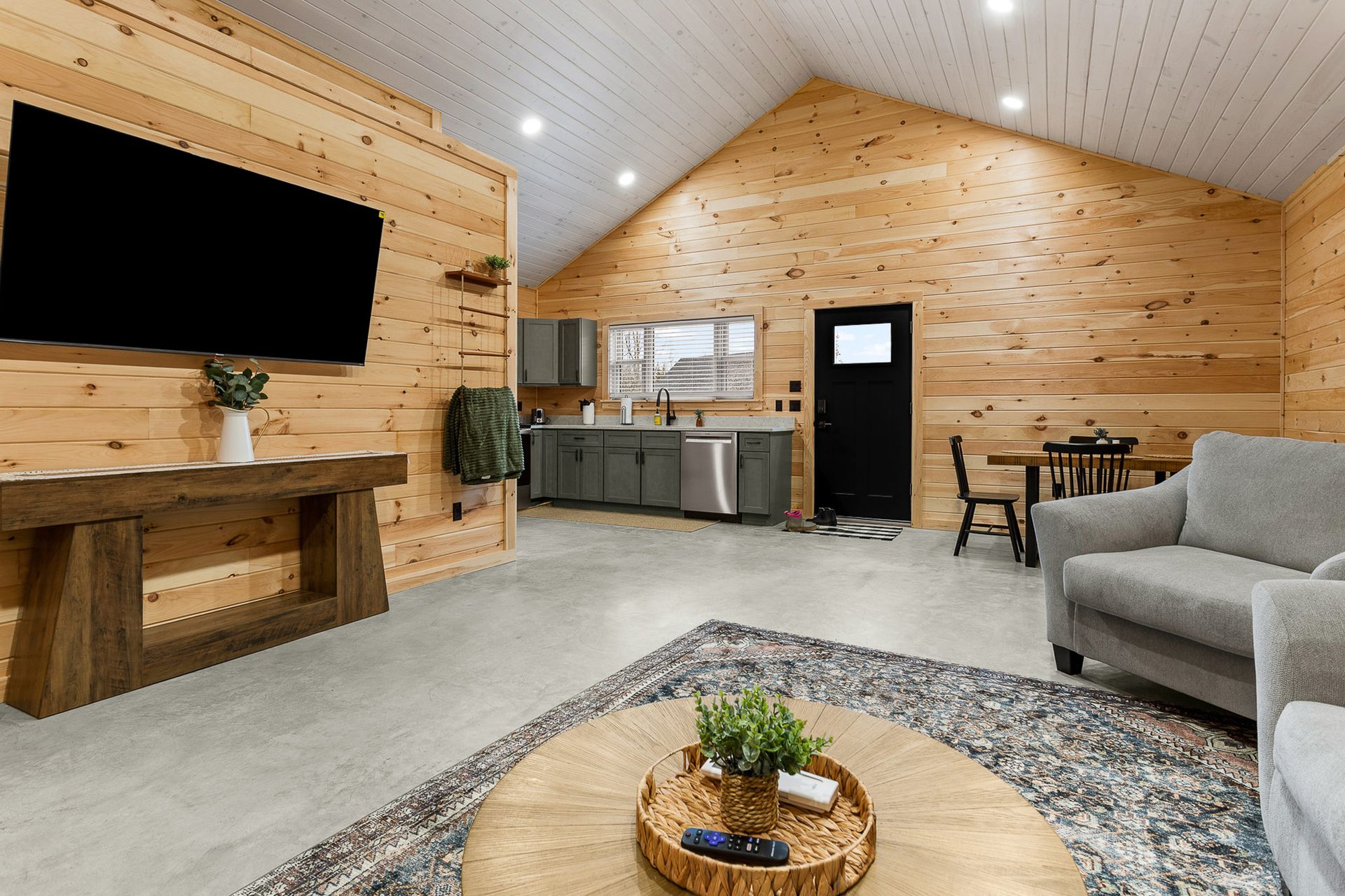 Modern living room with wood-paneled walls, wall-mounted TV, kitchen bar, gray sofa, and coffee table