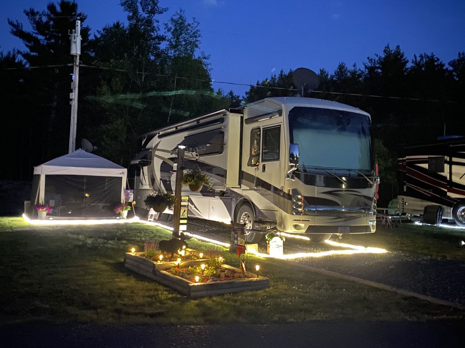 Lit RV parked at night beside a small illuminated flower bed and gazebo in a grassy yard.
