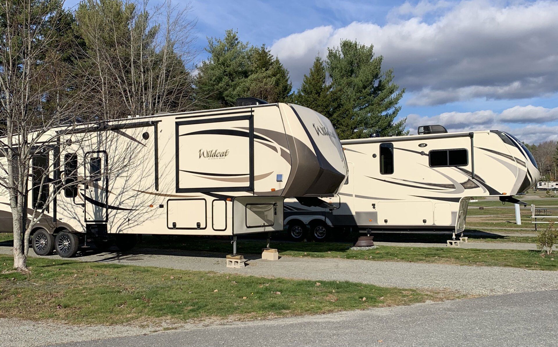 Two beige fifth-wheel campers parked on grass under a partly cloudy sky.