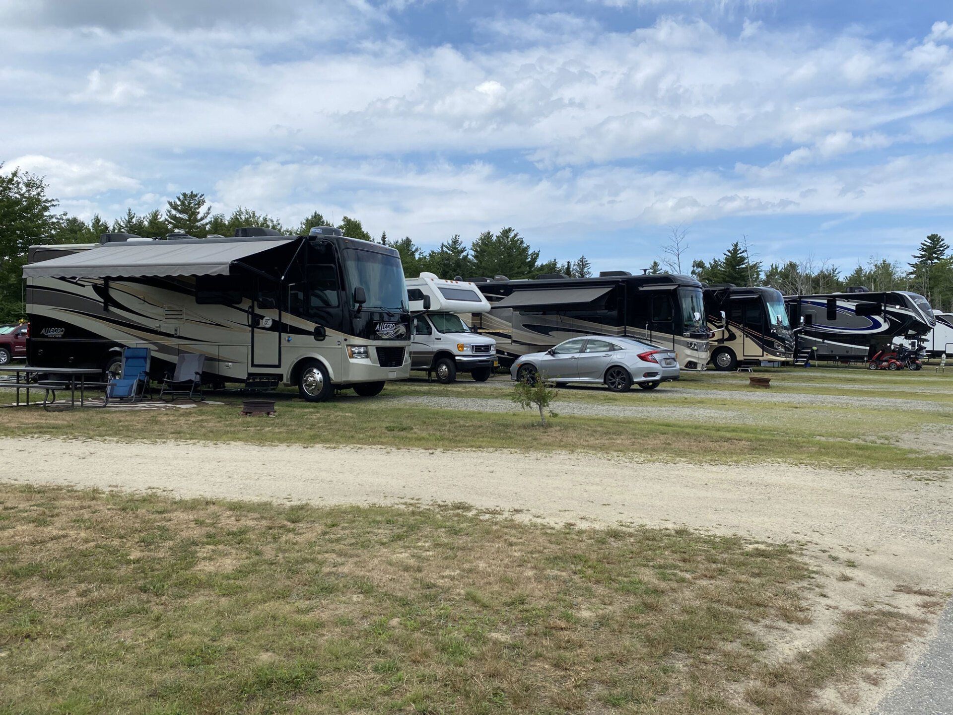 RVs and cars parked on a grassy lot under a partly cloudy sky, with trees in the background