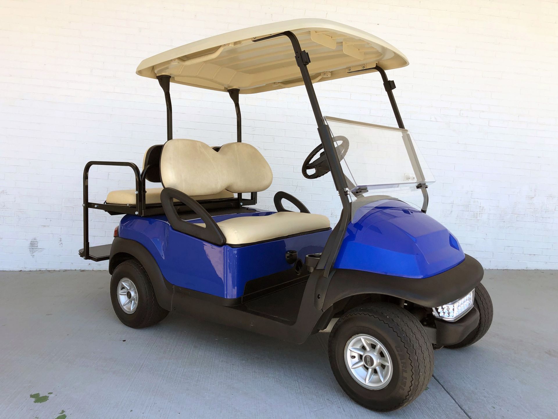 Blue golf cart with beige canopy and seats, parked on a paved surface against a white wall.