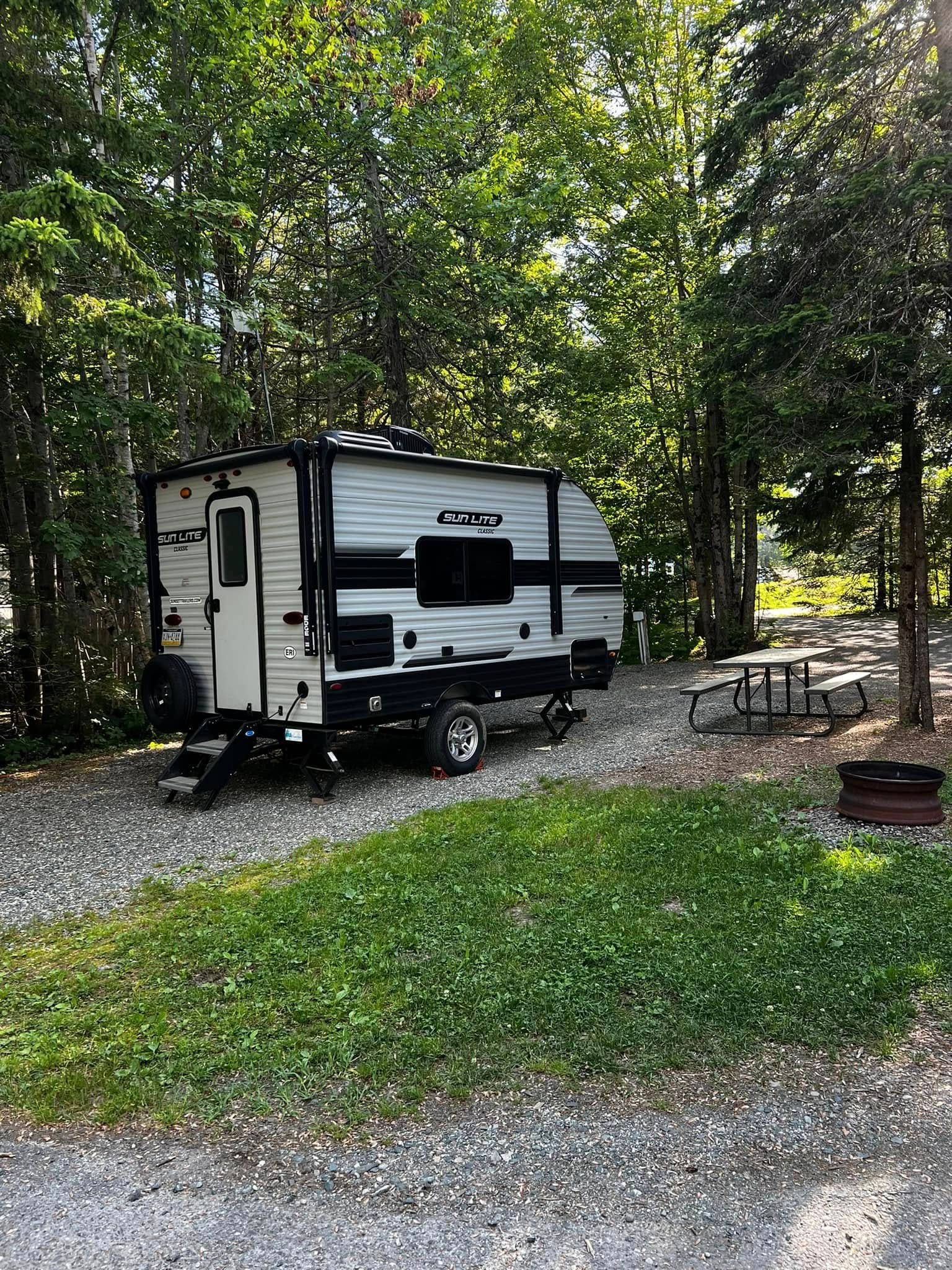 White travel trailer parked in a wooded campsite beside a picnic table and fire ring.