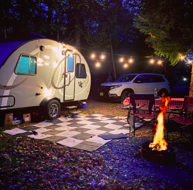 Cozy campsite with a small white camper, string lights, chairs, checkered patio, and a fire pit at night