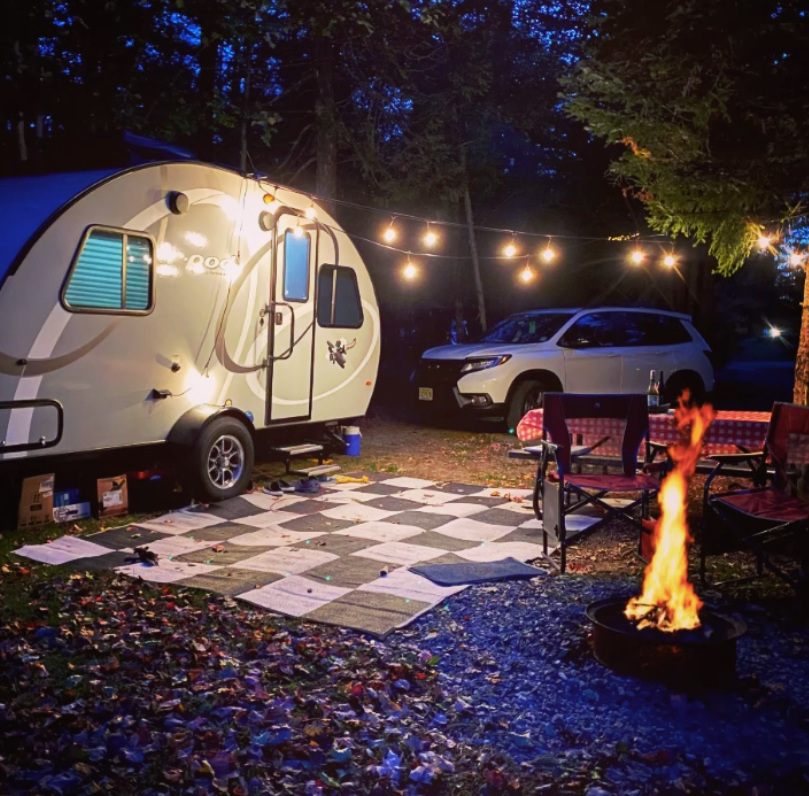 Nighttime campsite with a white teardrop trailer, string lights, picnic table, SUV, and campfire
