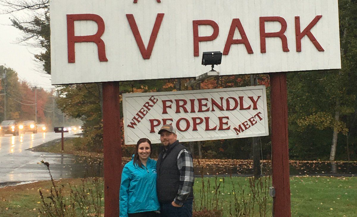 Two people stand beside an RV park sign that says “Friendly People Meet” by a wet roadside.