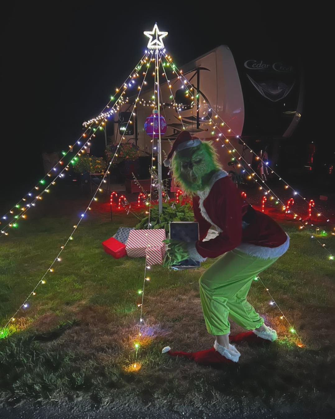 Person in green elf costume posing beside a lit Christmas tree and presents at night.
