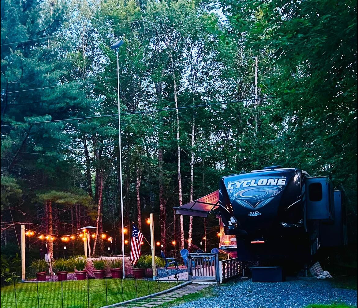 RV parked in a wooded campsite at dusk, with warm lights glowing among tall trees.