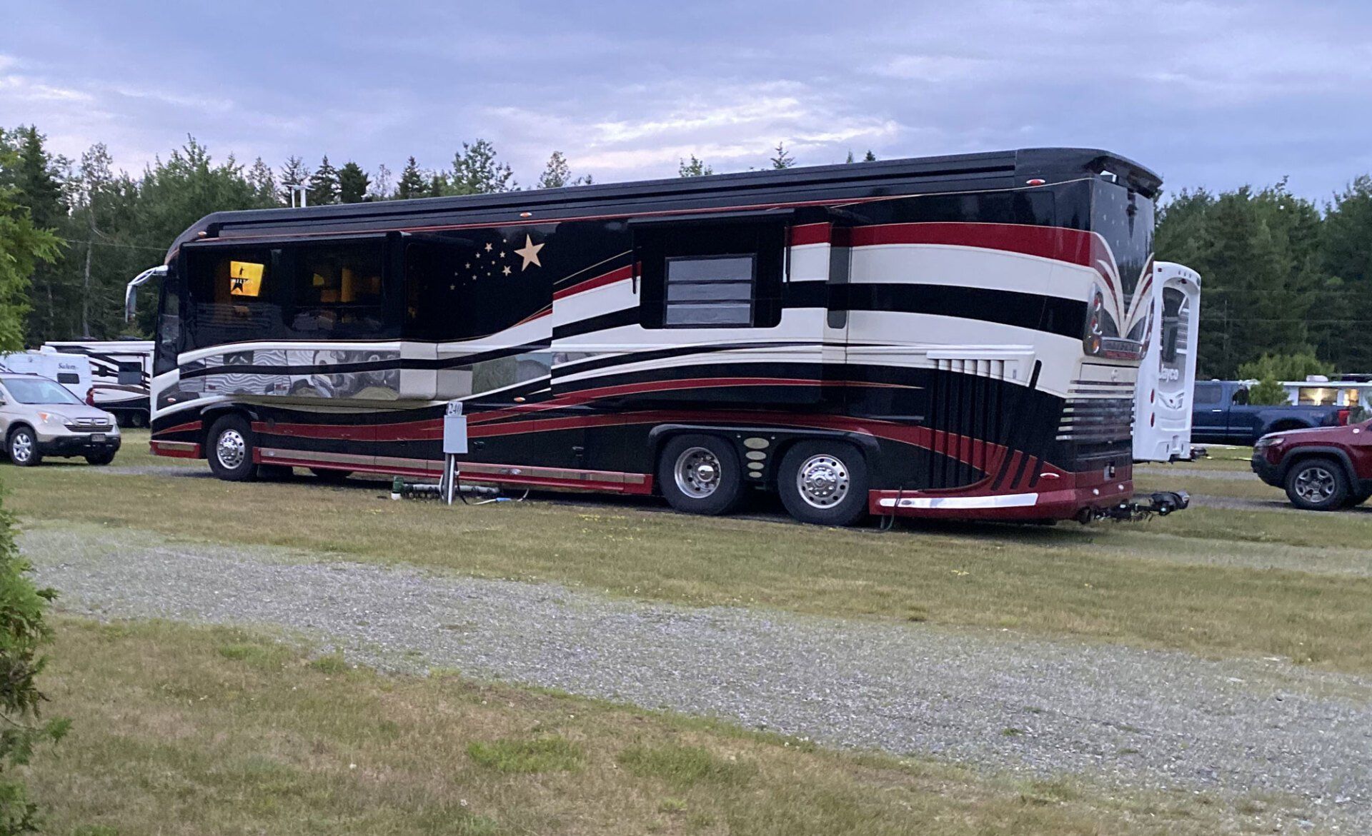 Large black-and-red bus parked on gravel near trees under a cloudy sky