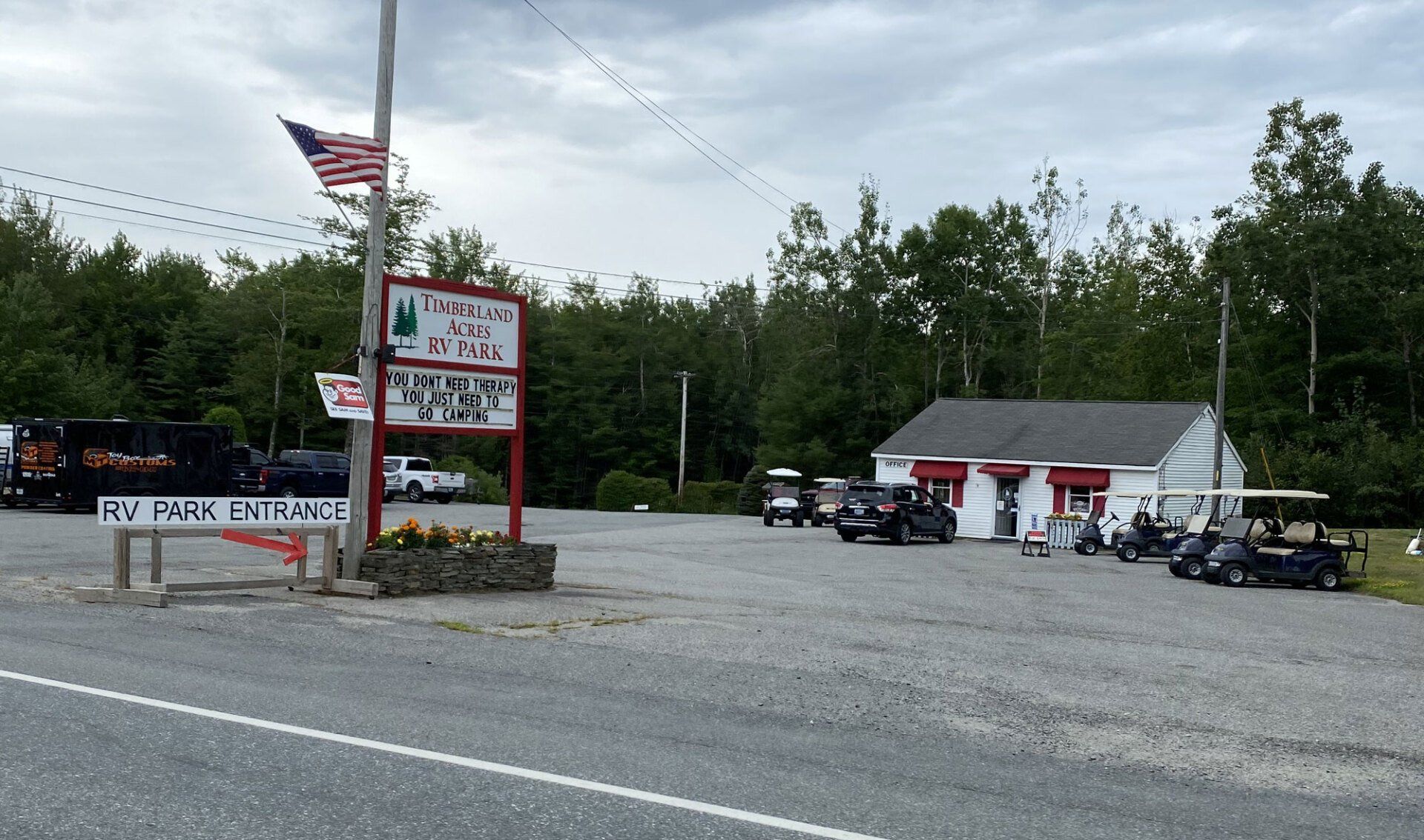 Gravel lot beside a roadside sign and American flag, with motorcycles parked near a small building and trees beyond.