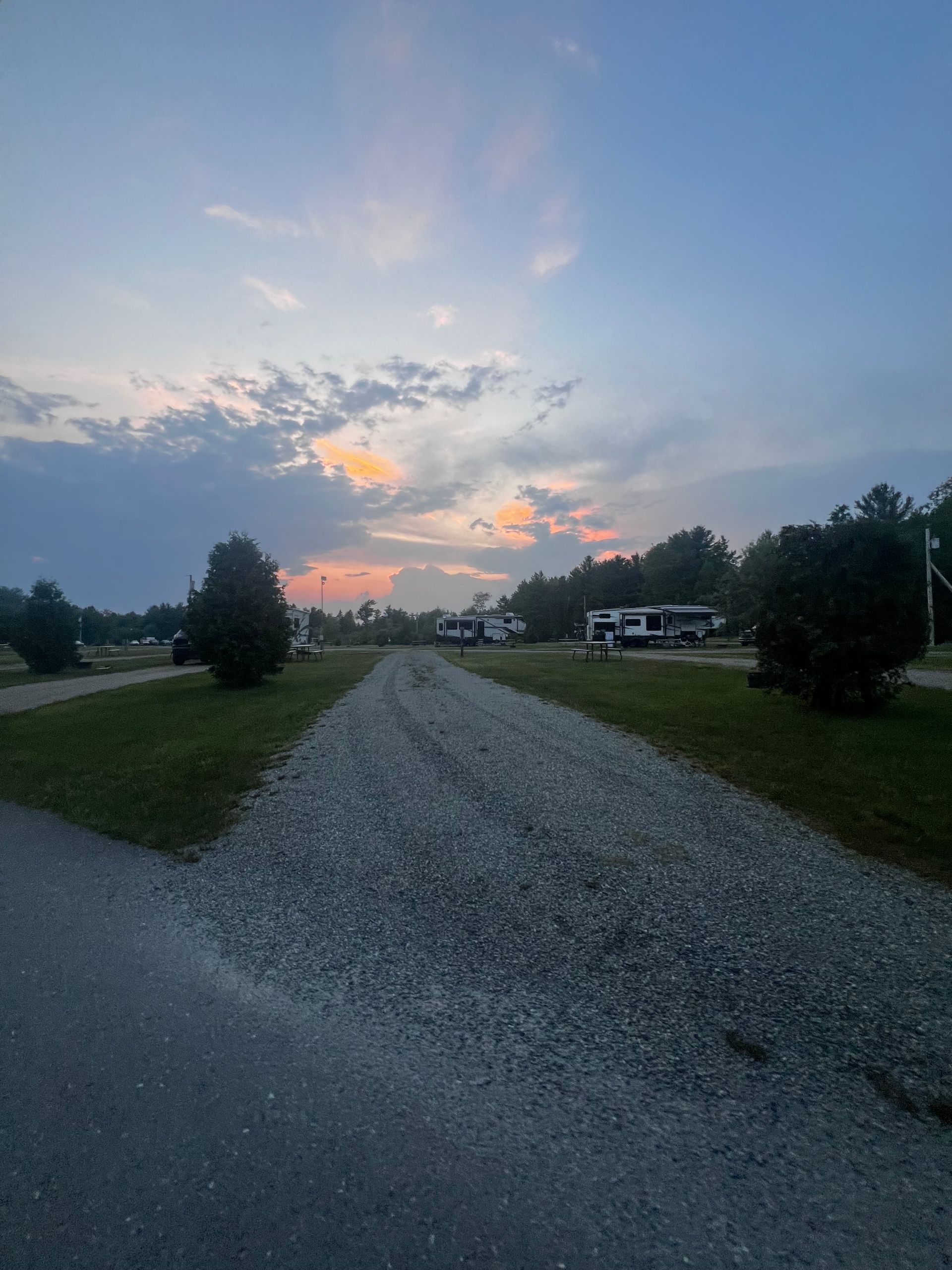 Gravel road leading toward houses at sunset, with green grass and a pastel sky