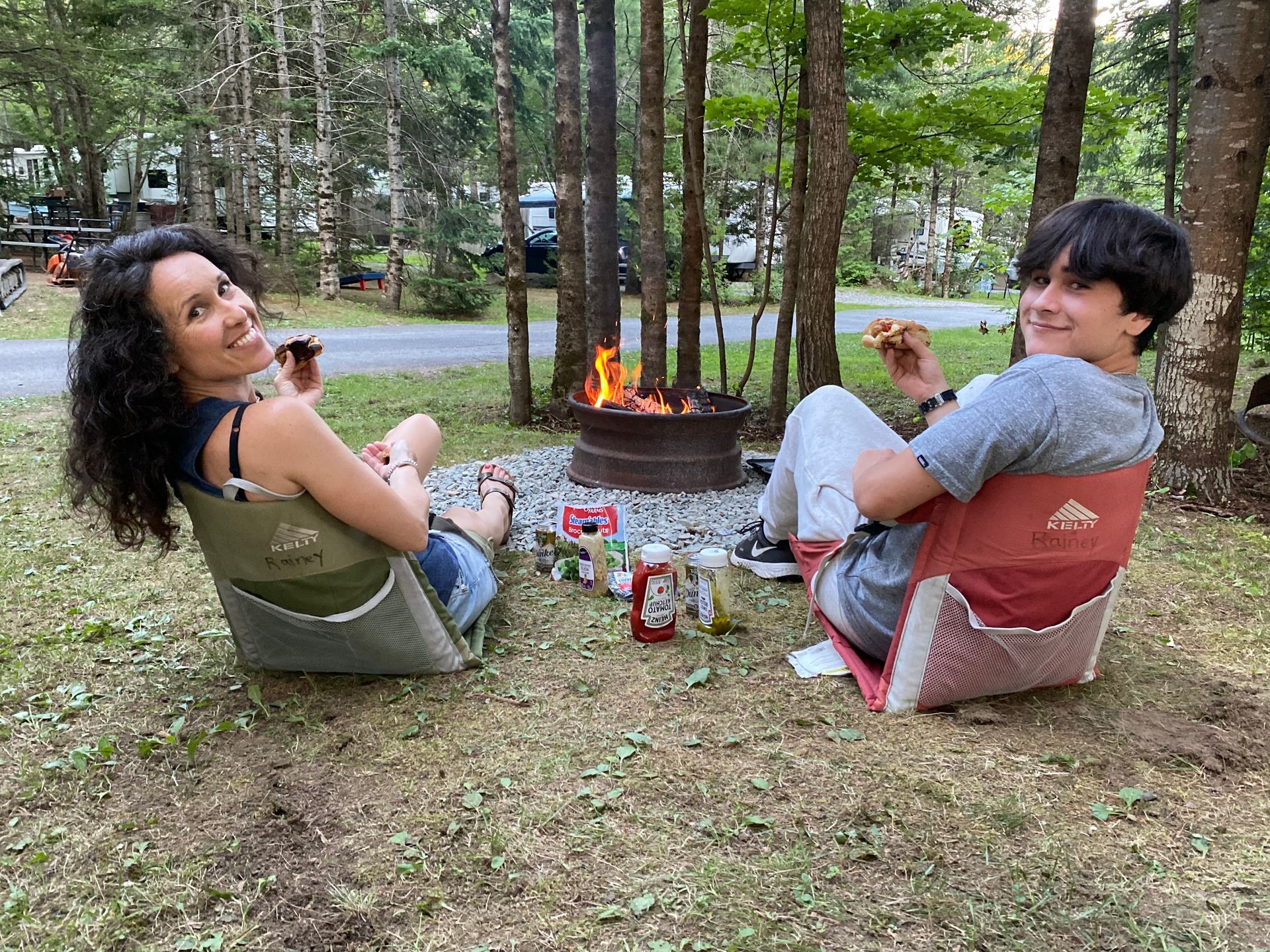 Two people relaxing by a campfire in a wooded campsite, smiling on folding chairs.