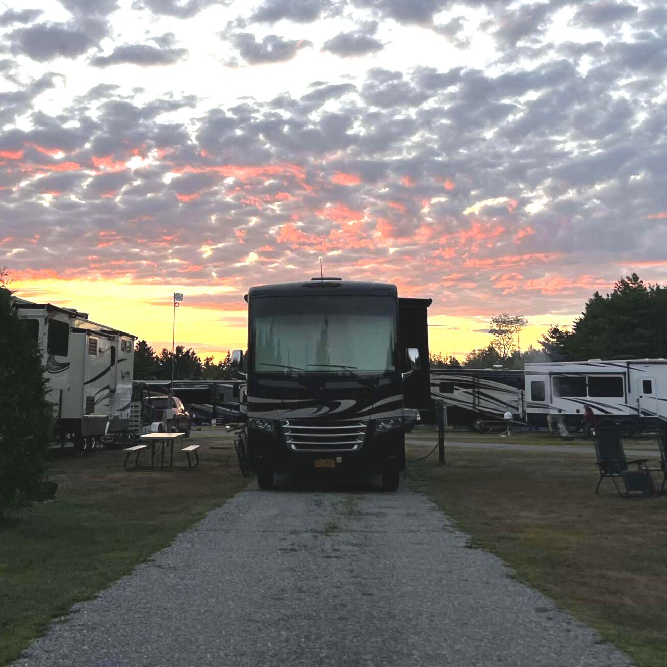 RV centered on a gravel path at sunset in a campground, framed by trailers and a colorful cloudy sky