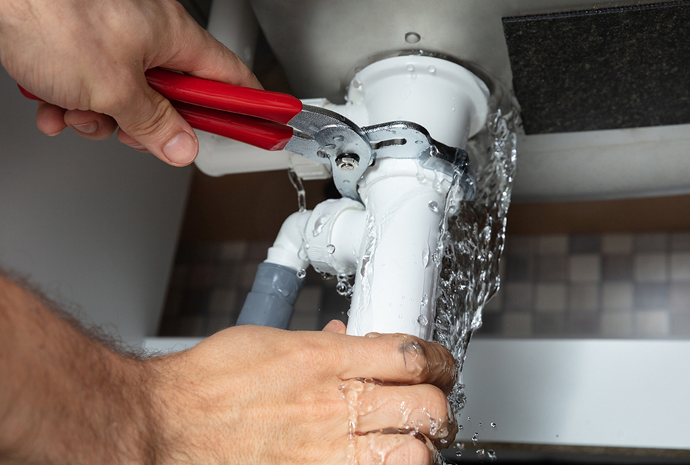 Hands using red pliers to fix a leaking white pipe under a sink