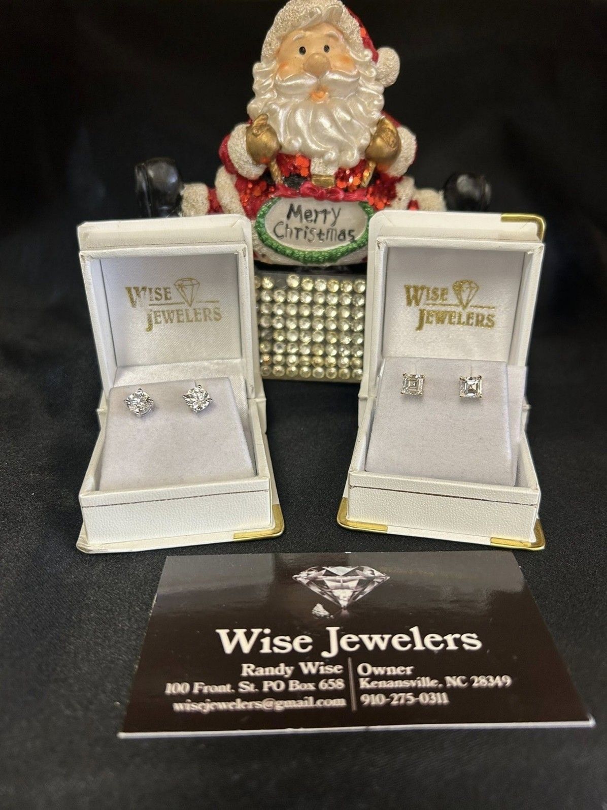 Two pairs of diamond earrings in open jewelry boxes with a Santa figure in the background. Wise Jewelers business card.