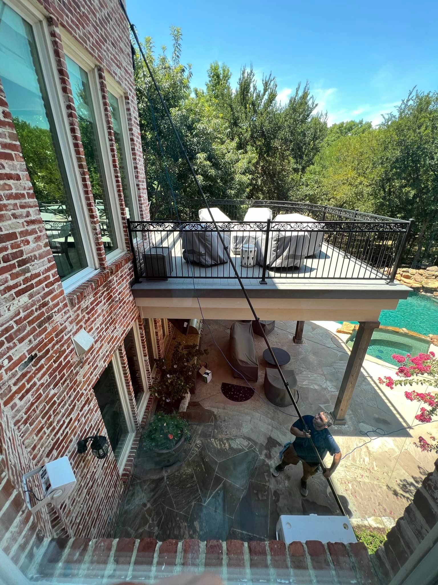 A person with a hose is spraying water on a patio under a deck, near a pool and red brick building on a sunny day.