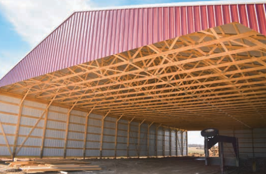Open view inside a red-roofed barn with exposed wooden trusses and side walls, with light streaming through the open end.