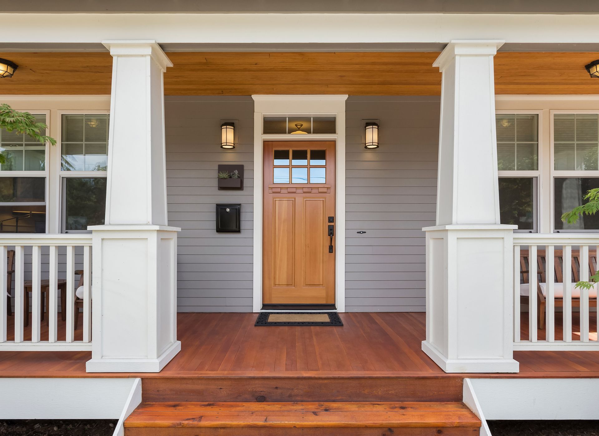 Front porch of a gray house with a wooden door, white columns, and railing; warm wooden deck.