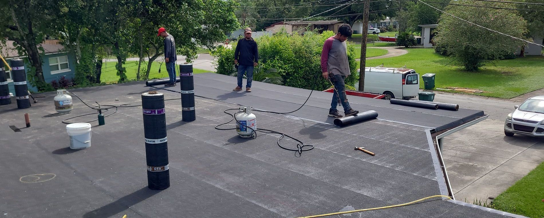 Workers on a flat roof installing roofing materials. Propane tanks and tools are visible.