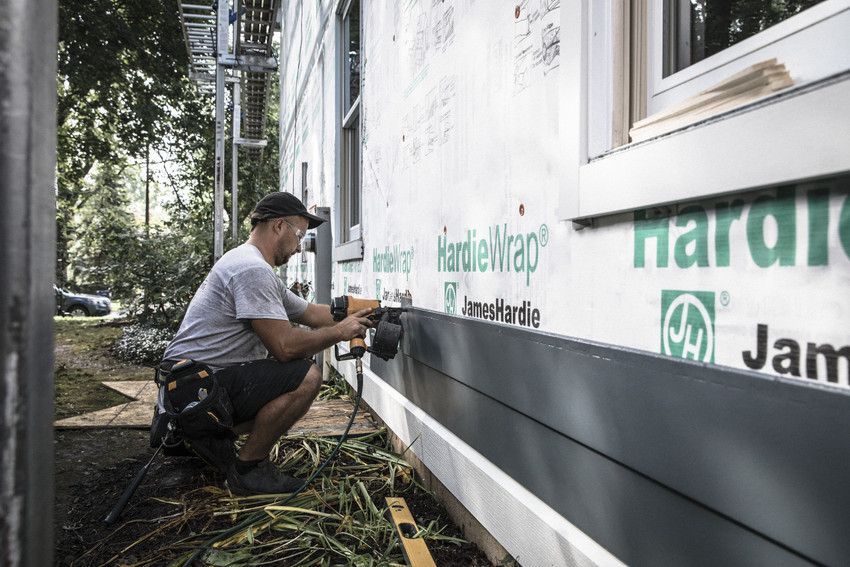 A construction worker installs dark gray siding on a house, using a nail gun.