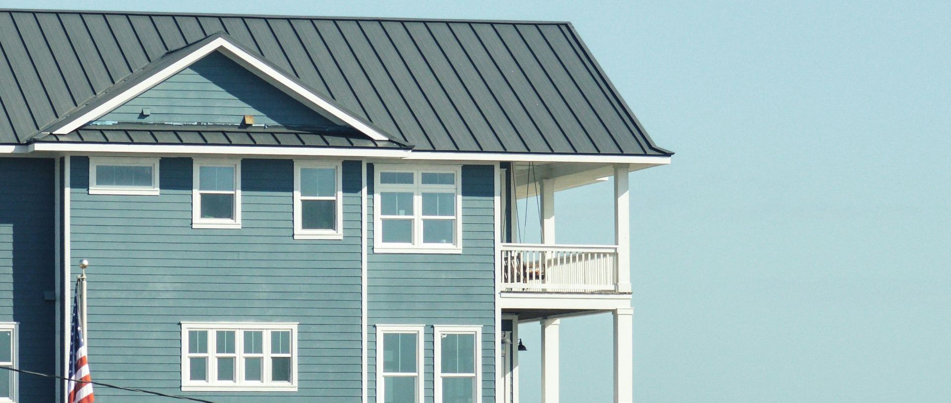 Blue house with white trim and a porch, against a light blue sky. The house has a dark gray metal roof.