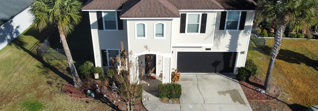 Two-story beige house with black shutters, a driveway, and a garage. Palm trees flank the front.
