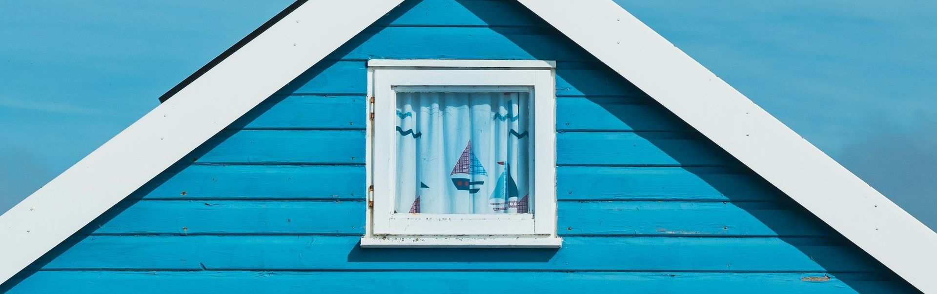 Blue beach hut with white trim and a window, showcasing miniature sailboats inside.