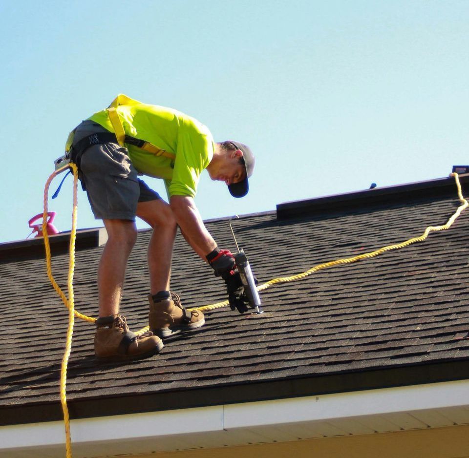 A roofer in a safety harness on a roof repairs shingles using a tool. The sky is clear blue.