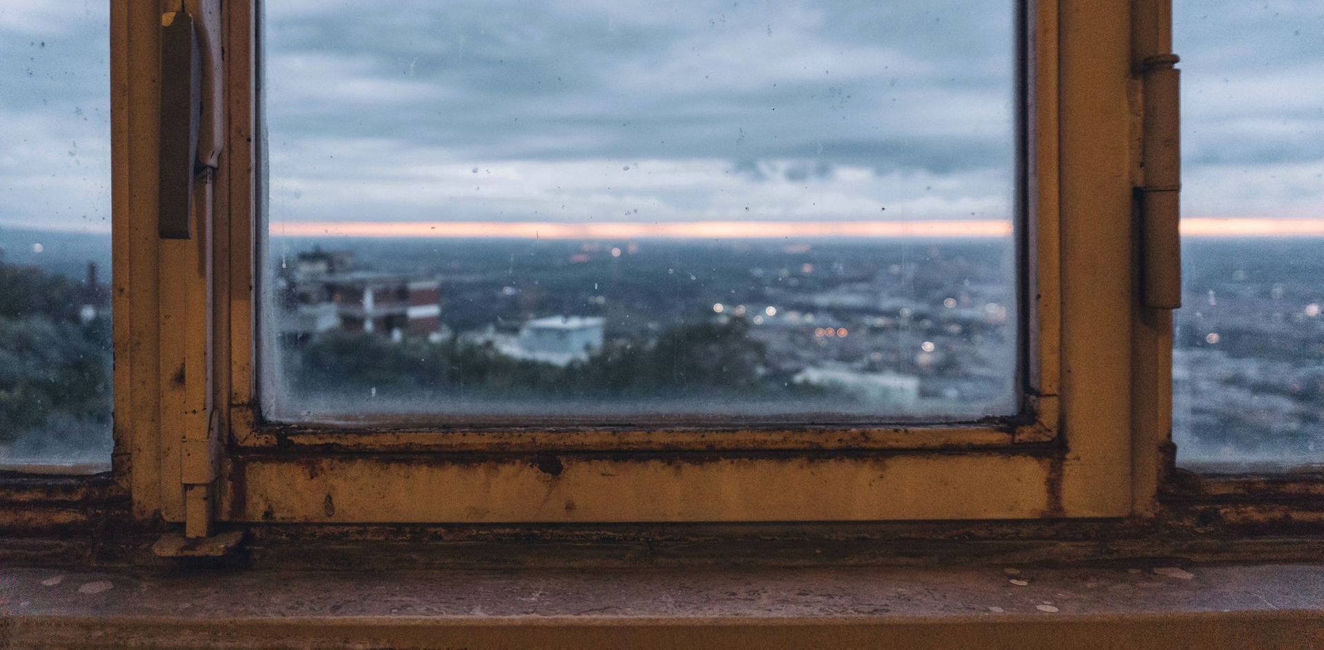 View of a cityscape through a weathered window. The sky is overcast, with a pale band of light on the horizon.