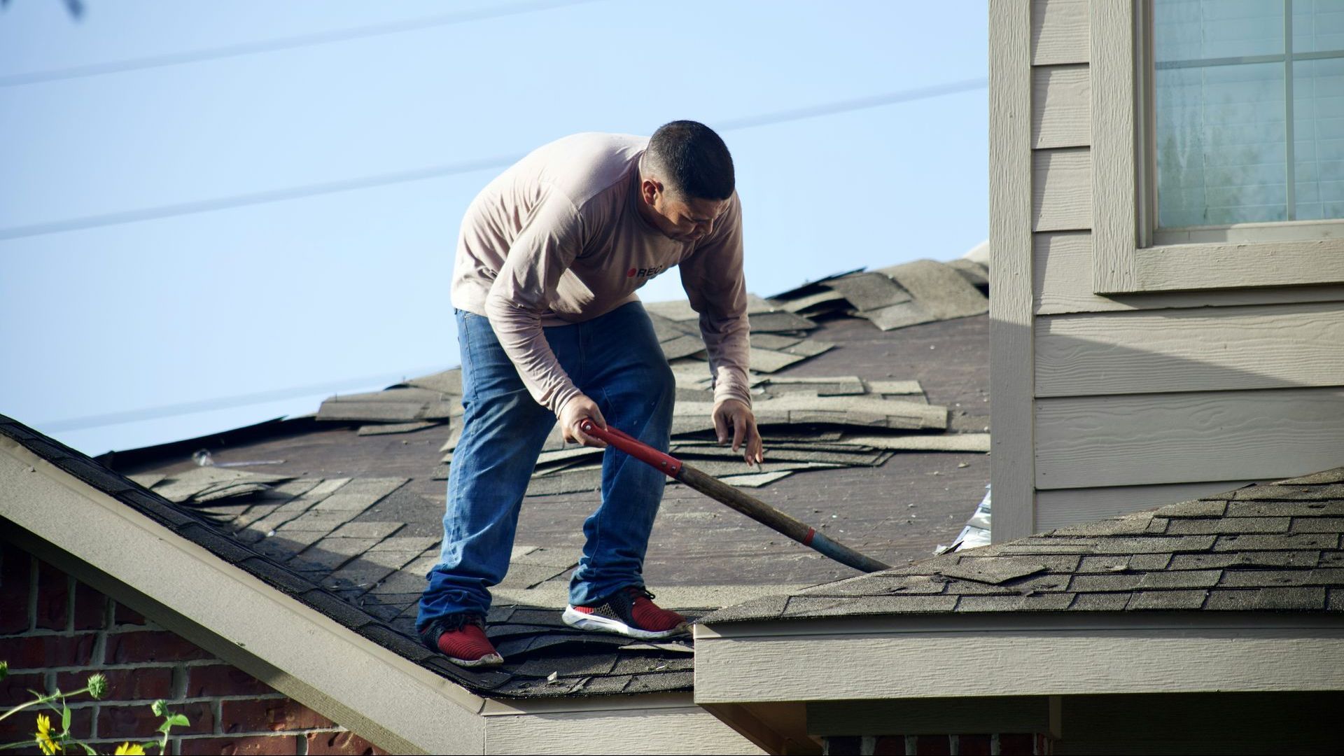 Man in blue jeans and a tan shirt removing damaged shingles from a house roof with a tool.