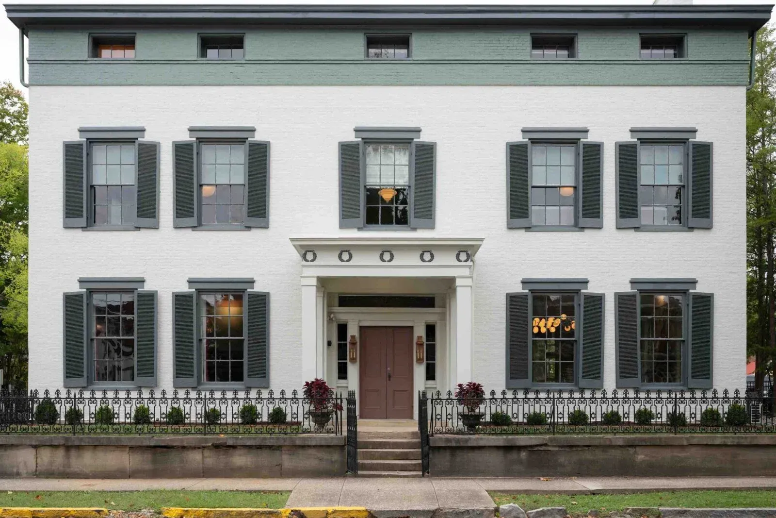 Front view of a white two-story house with green shutters, a central porch, and black iron fence.