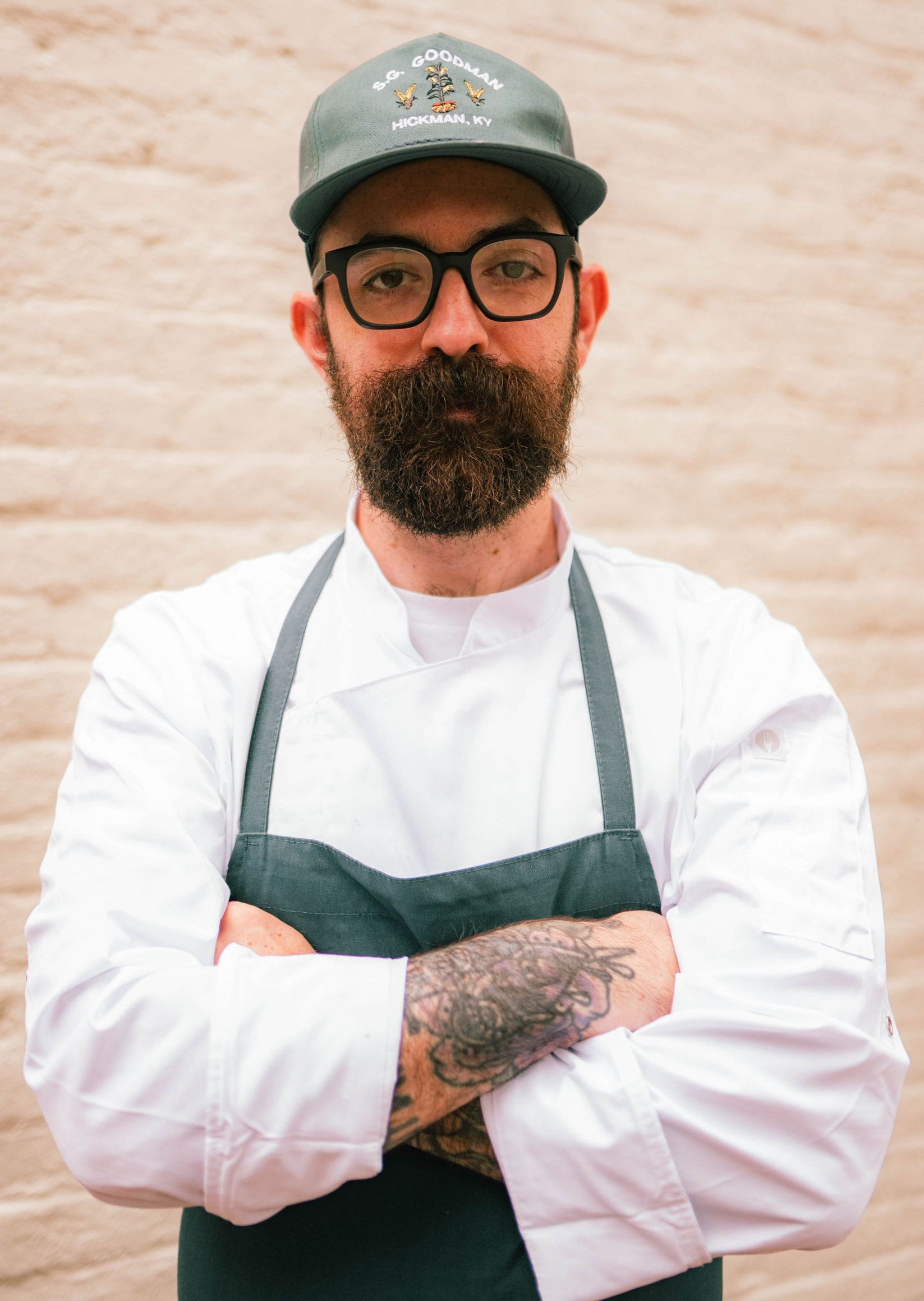 Chef in a white coat and apron, arms crossed, wearing glasses and a cap, standing against a brick wall