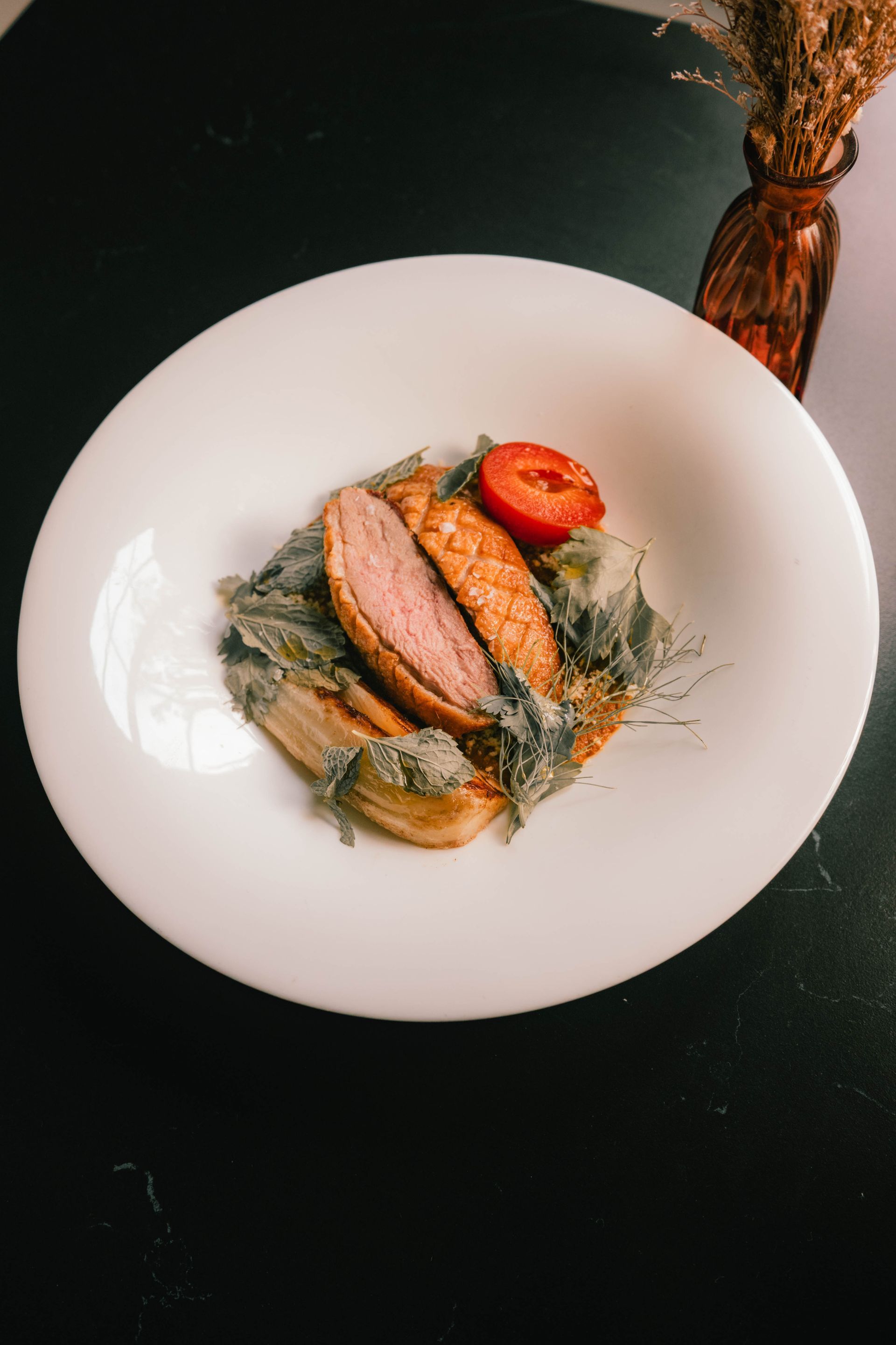 Plated gourmet dish with sliced meat, tomato, and greens on a white plate against a dark background