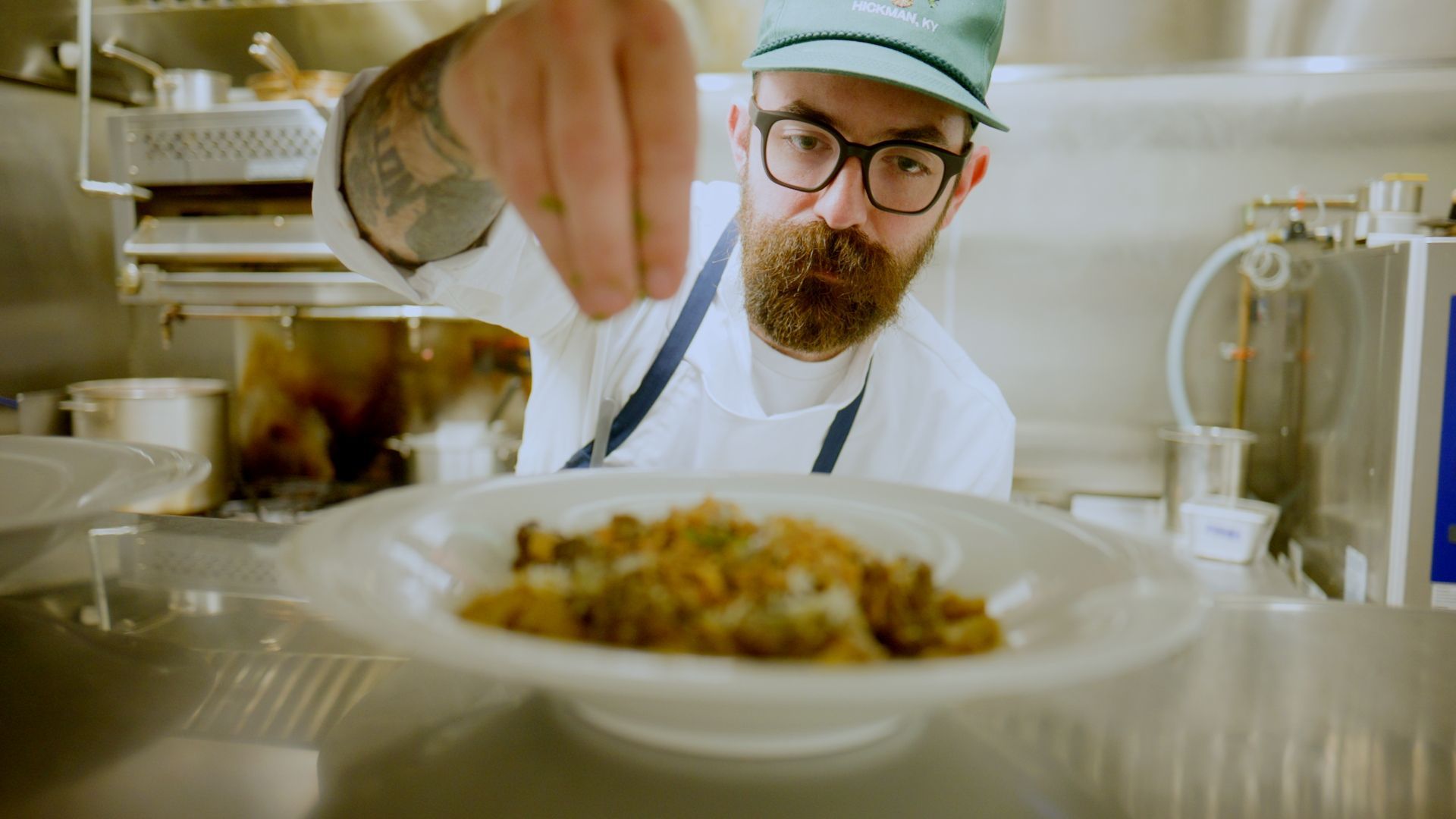 Chef in white coat and green cap reaching toward a plated dish in a bright kitchen.
