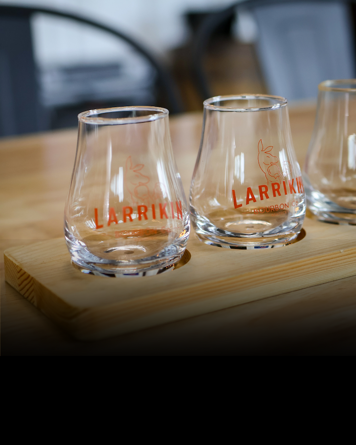 Three clear branded glasses on a wooden tray, one partially filled with amber liquid