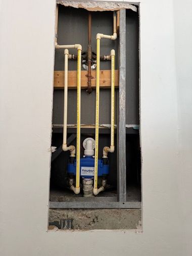 Water heater plumbing with red and blue shutoff valves and flexible braided hoses above a dusty floor.
