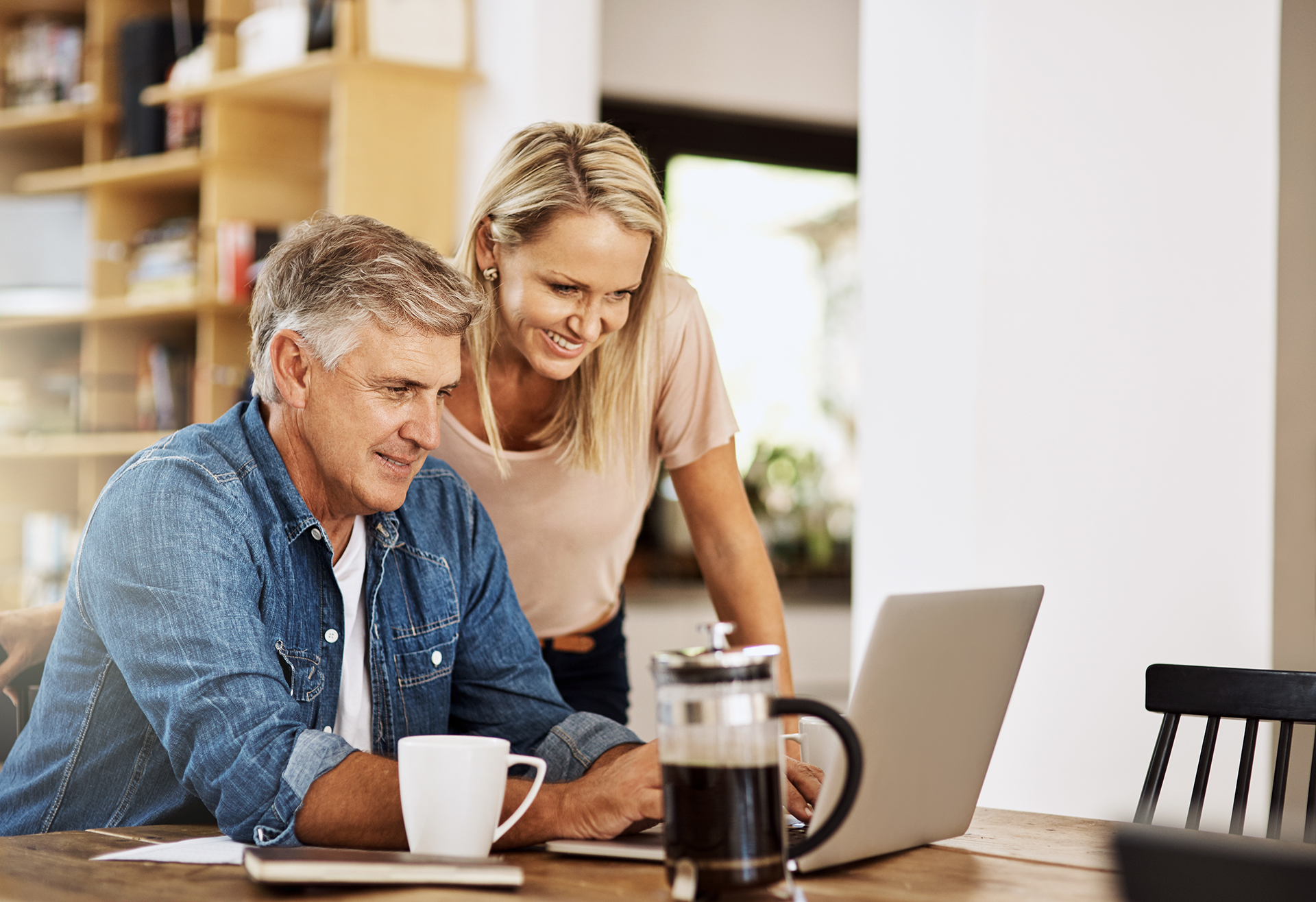 Two people smiling at a laptop on a kitchen table with coffee mugs and a French press