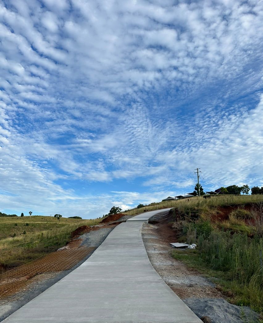 A Concrete Road Going Through A Field With A Blue Sky And Clouds — Hinterland Concreting Pty Ltd In Lennox Head, NSW