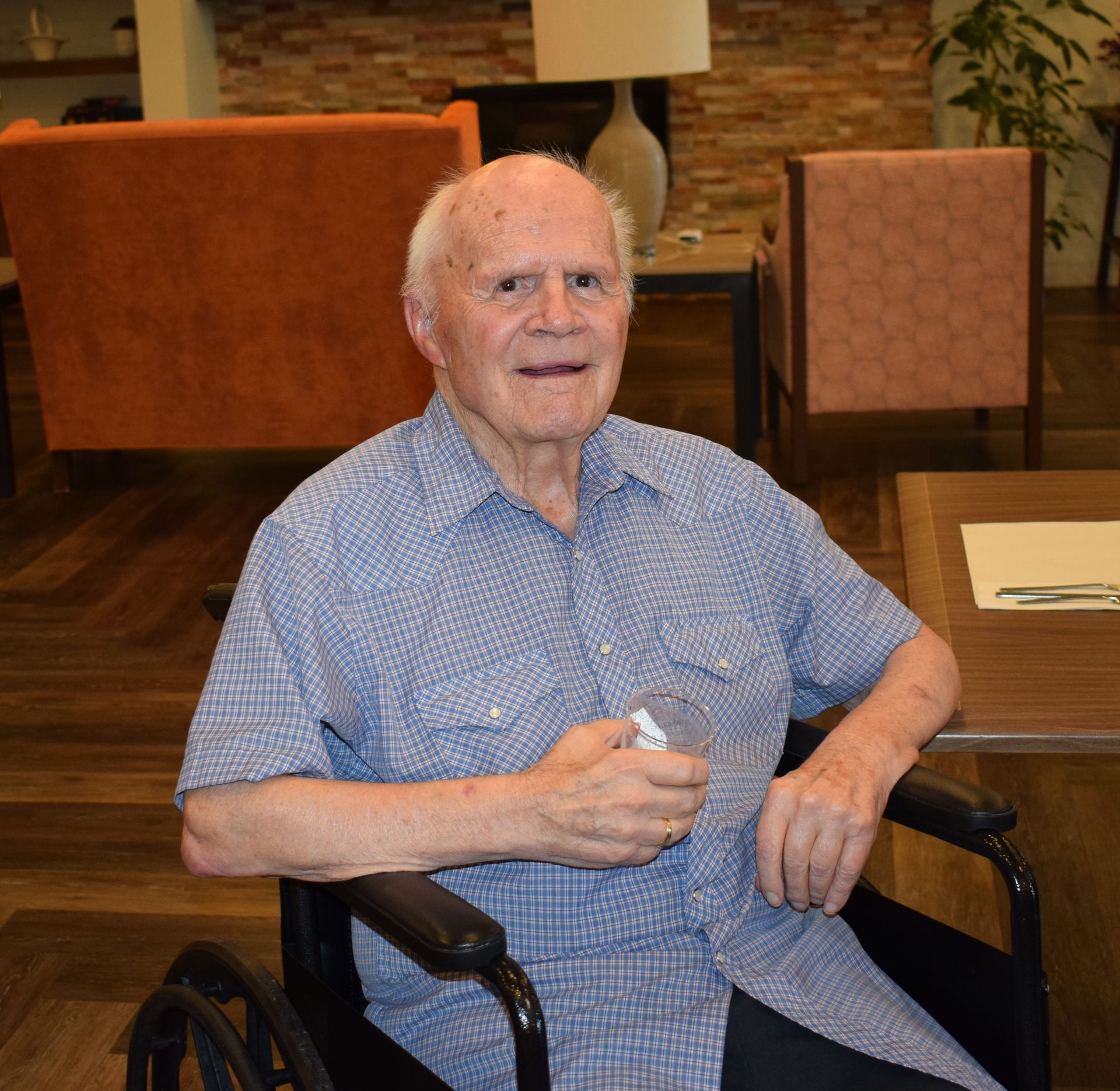 A person in a blue button-down shirt sitting in a wheelchair indoors, holding a small clear glass.