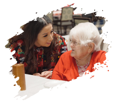 Young woman smiles at an elderly woman in a red sweater. They sit at a table indoors.