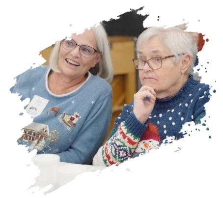 Two older women in holiday sweaters, one smiling and the other deep in thought, sitting together.