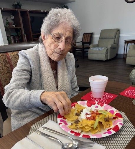 An older adult with short, light hair eats a plate of nachos at a table with a white cup and silverware nearby.