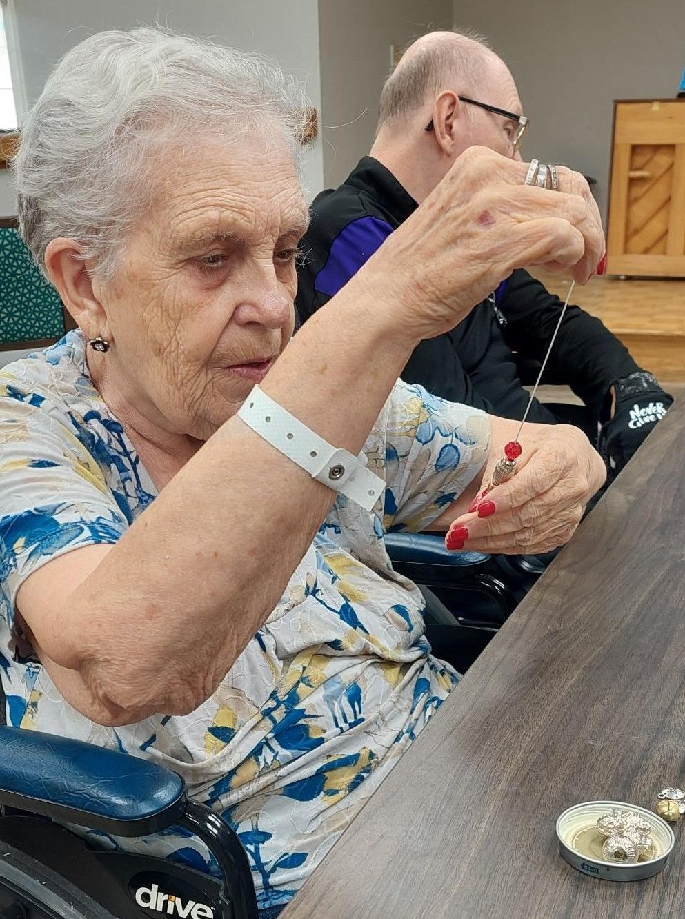 A person in a wheelchair, wearing a medical ID bracelet, assembles jewelry at a table with another person in the background.
