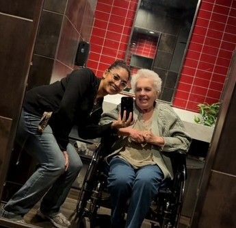 A person stands smiling next to someone in a wheelchair inside a bathroom with red-tiled walls, reflected in a mirror.