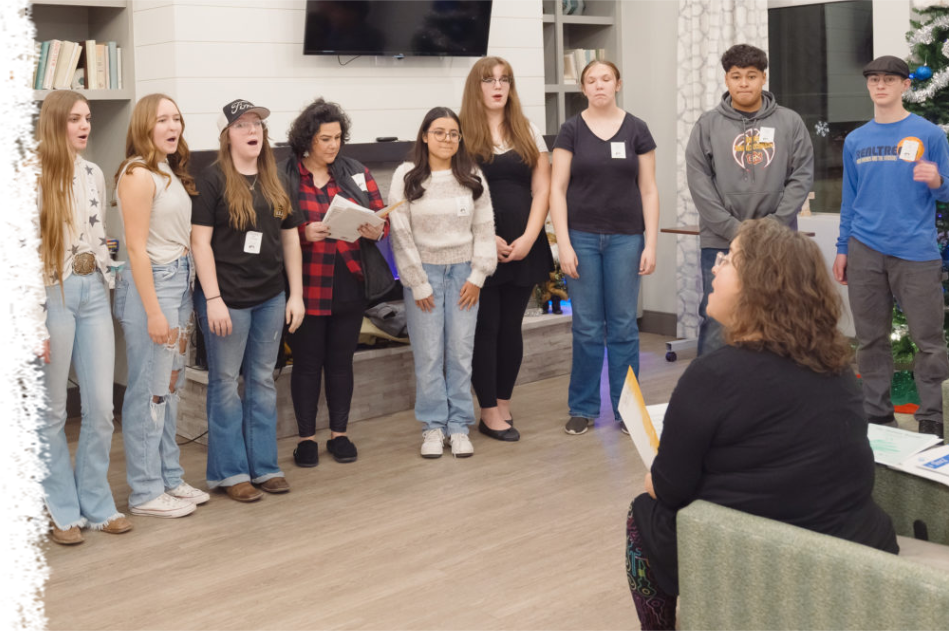 A choir stands in a living room, singing while an instructor in the foreground leads them from an armchair.